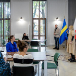 Pani p.o. Zastępcy Dyrektora Biura Spraw Społecznych Monika Siemion przemawia do grupy siedzącej przy stołach w sali z flagami województwa śląskiego, Polski i Unii Europejskiej.