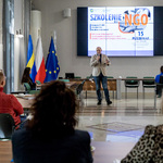 Pan Tomasz Pawłowski wita uczestników szkolenia dla NGO w sali z ekranem, na którym wyświetlony jest tytuł wydarzenia, oraz flagami województwa śląskiego, Polski i Unii Europejskiej