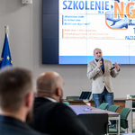 Pan Tomasz Pawłowski przemawia do uczestników szkolenia dla NGO w sali z ekranem, na którym wyświetlony jest tytuł wydarzenia, oraz flagami województwa śląskiego, Polski i Unii Europejskiej