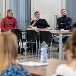 Grupa osób siedzi przy stołach w sali konferencyjnej podczas dyskusji, jedna osoba trzyma mikrofon i notatnik.