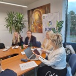 Fragment stołu konferencyjnego z laptopem, dokumentami i napojem; w tle baner RDPP oraz obraz na ścianie.