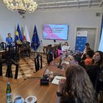 Uczestnicy spotkania siedzą przy stołach ustawionych w literę U. W tle ekran ze slajdem początkowym.