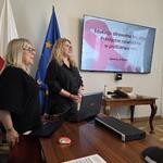 Dwie kobiety stoją i przemawiają do uczestników warsztatów, w tle ekran ze slajdem tytułowym.
