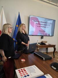 Dwie kobiety stoją przy stole podczas prezentacji; na ekranie widoczny slajd o edukacji zdrowotnej i profilaktyce HIV